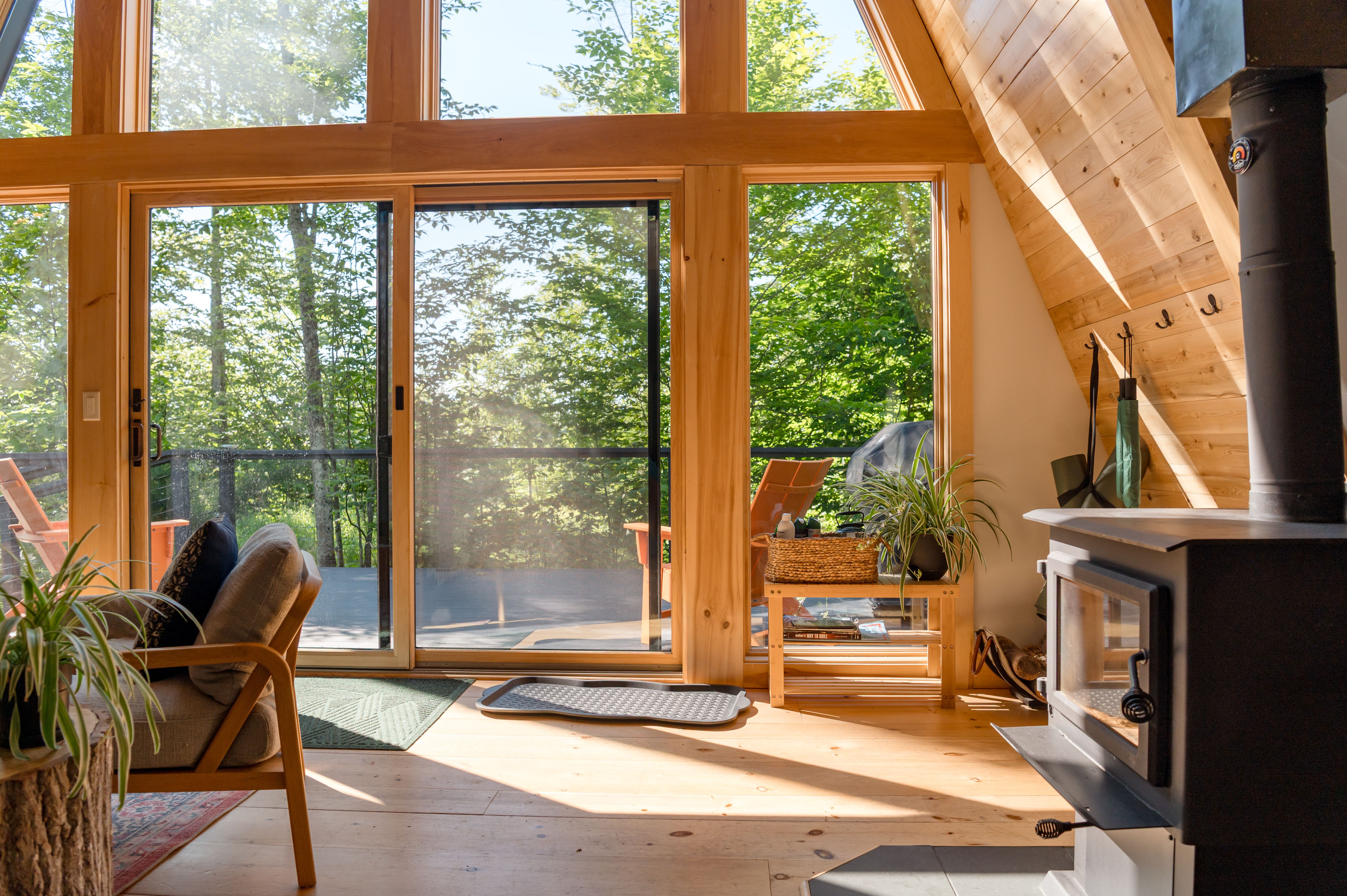 Sunlit interior of a cozy A-frame cabin with a wood stove, overlooking a green deck.