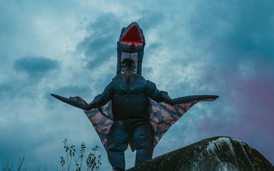 A person in a dinosaur costume poses under a dramatic, cloudy sky in Uribelarrea.