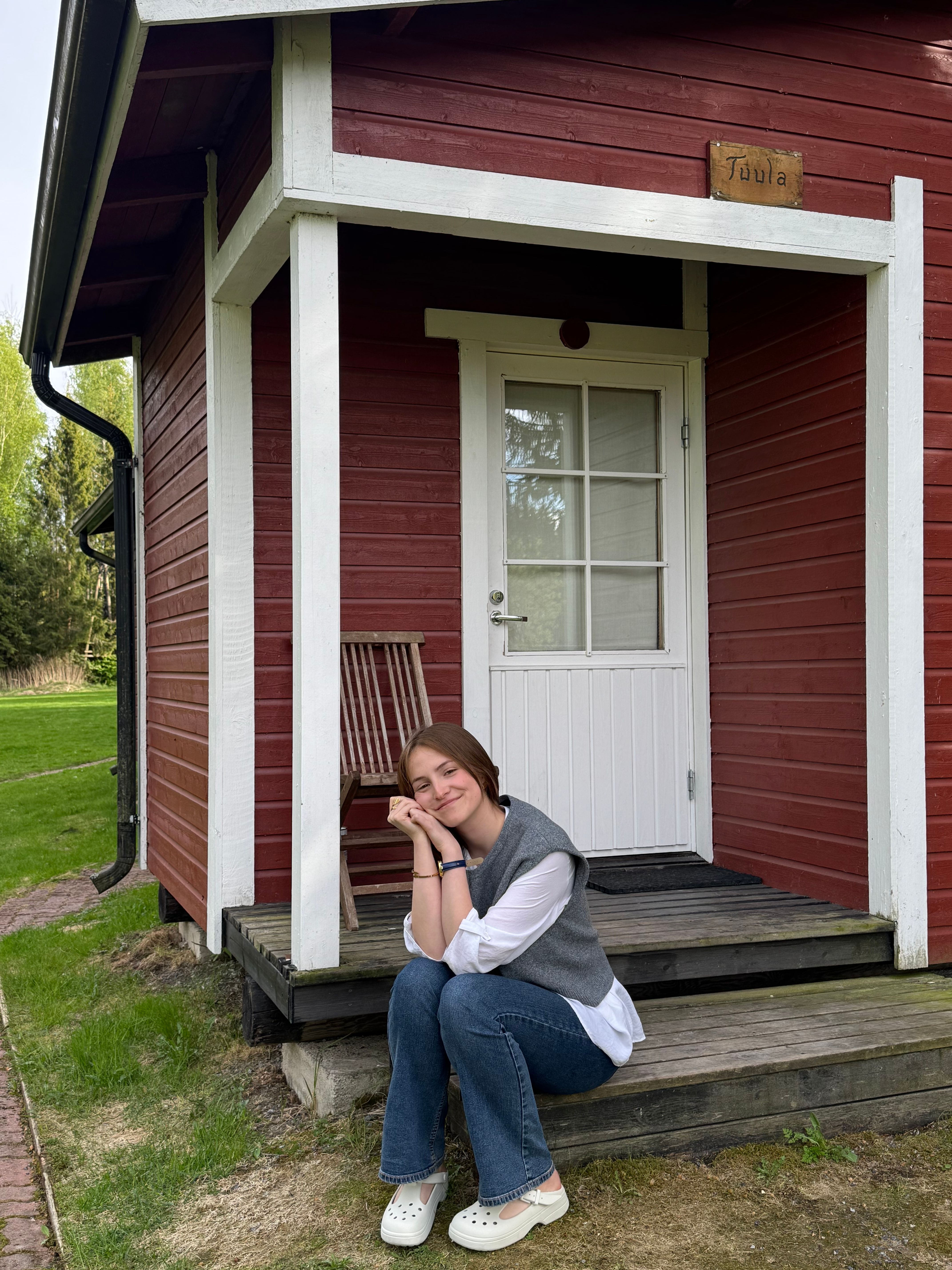 A relaxed young woman poses on the steps of a charming red cabin in a serene outdoor setting.