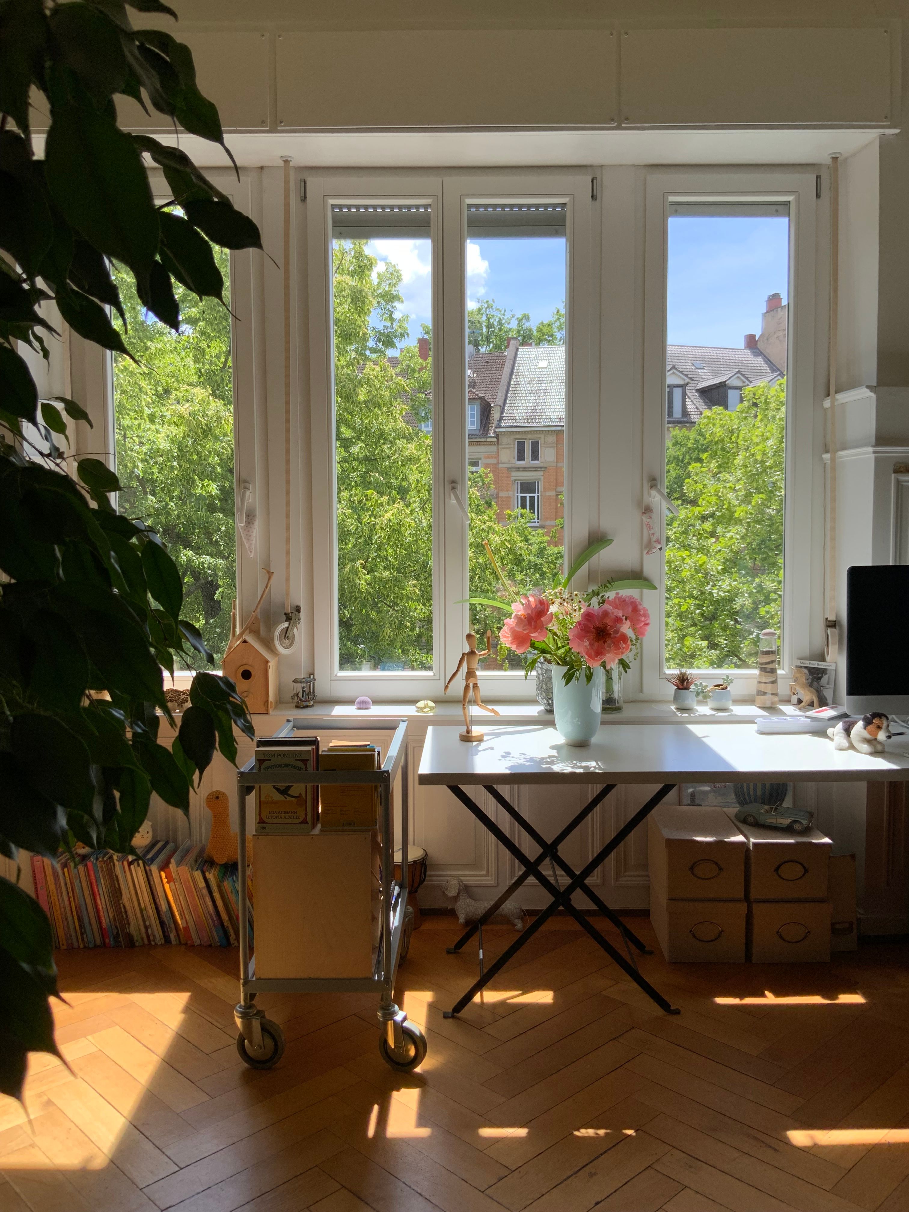 A sunny home office with a desk, plants, and large windows overlooking trees.