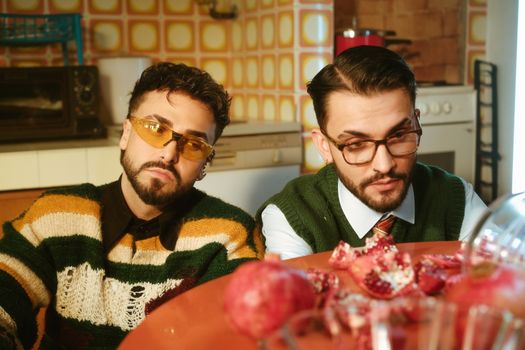 Two stylish men wearing glasses pose in a retro kitchen with pomegranates scattered on the table.