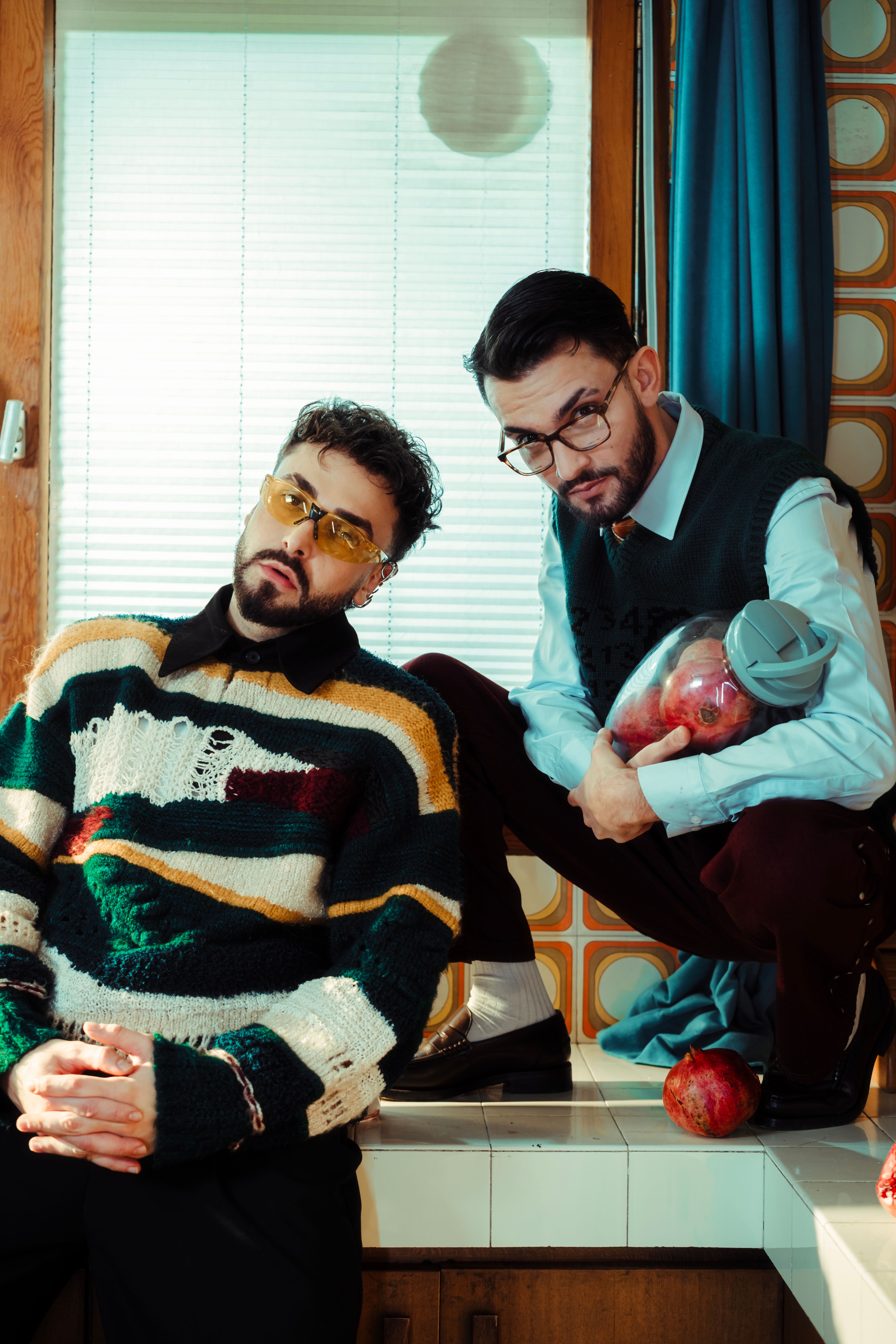 Two stylish men in retro fashion pose creatively with pomegranates indoors.