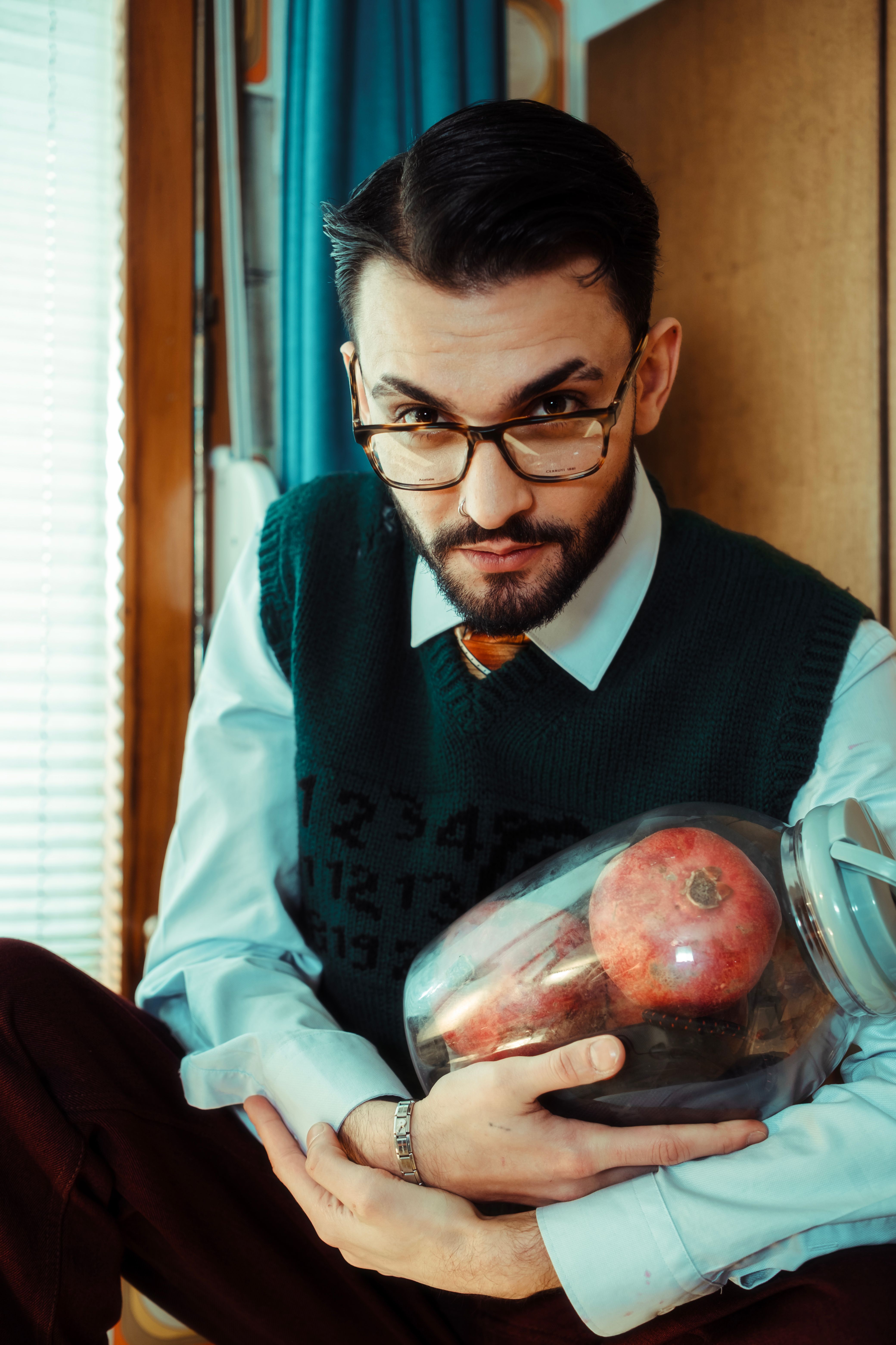 Young man in glasses holding jar with pomegranates indoors, thoughtful expression.