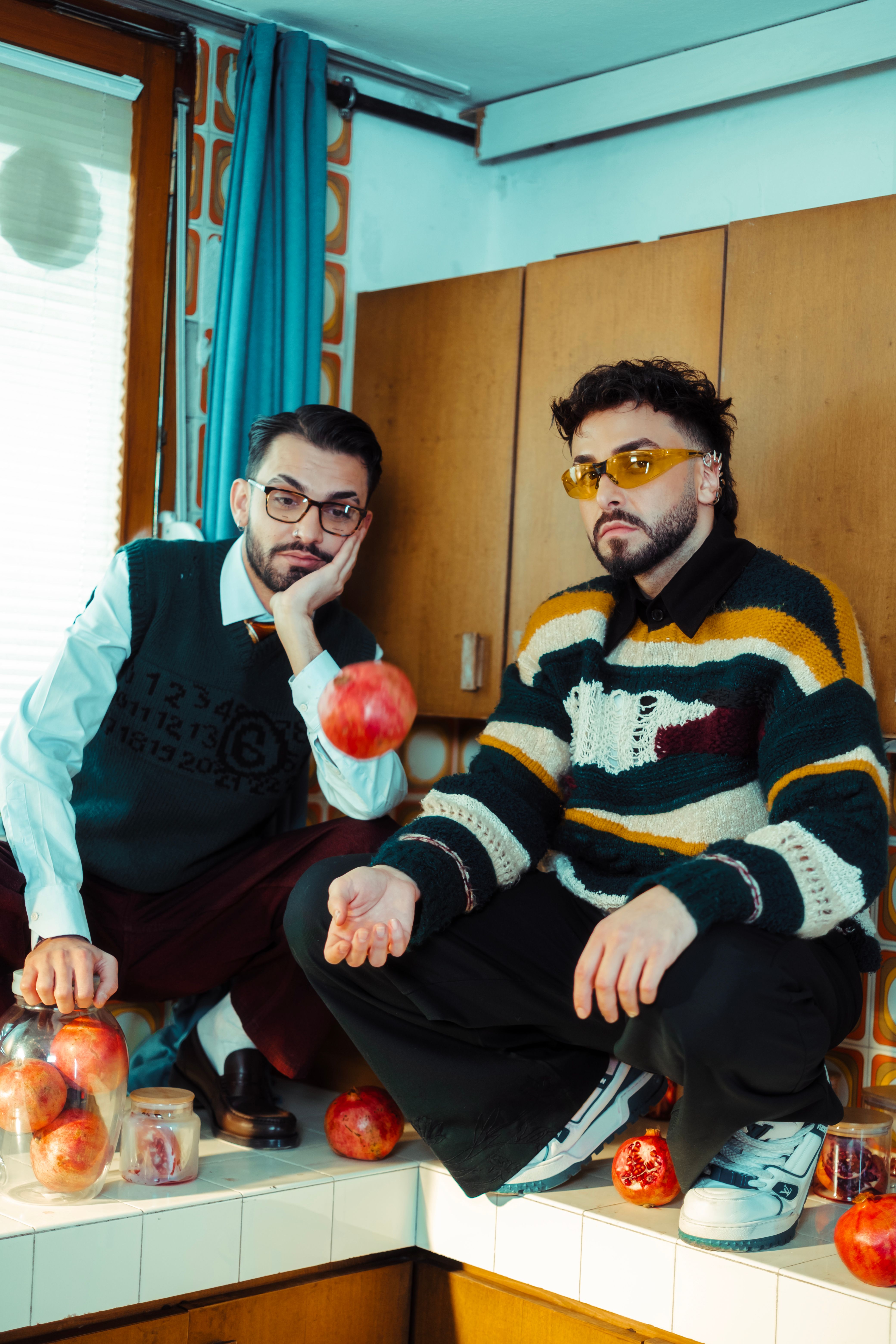 Two men in retro attire sit in a vintage kitchen surrounded by apples.