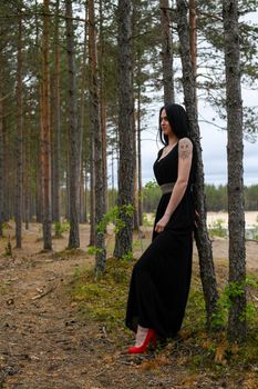 A fashionable woman in a black dress with red shoes leans against a tree in an Estonian forest.