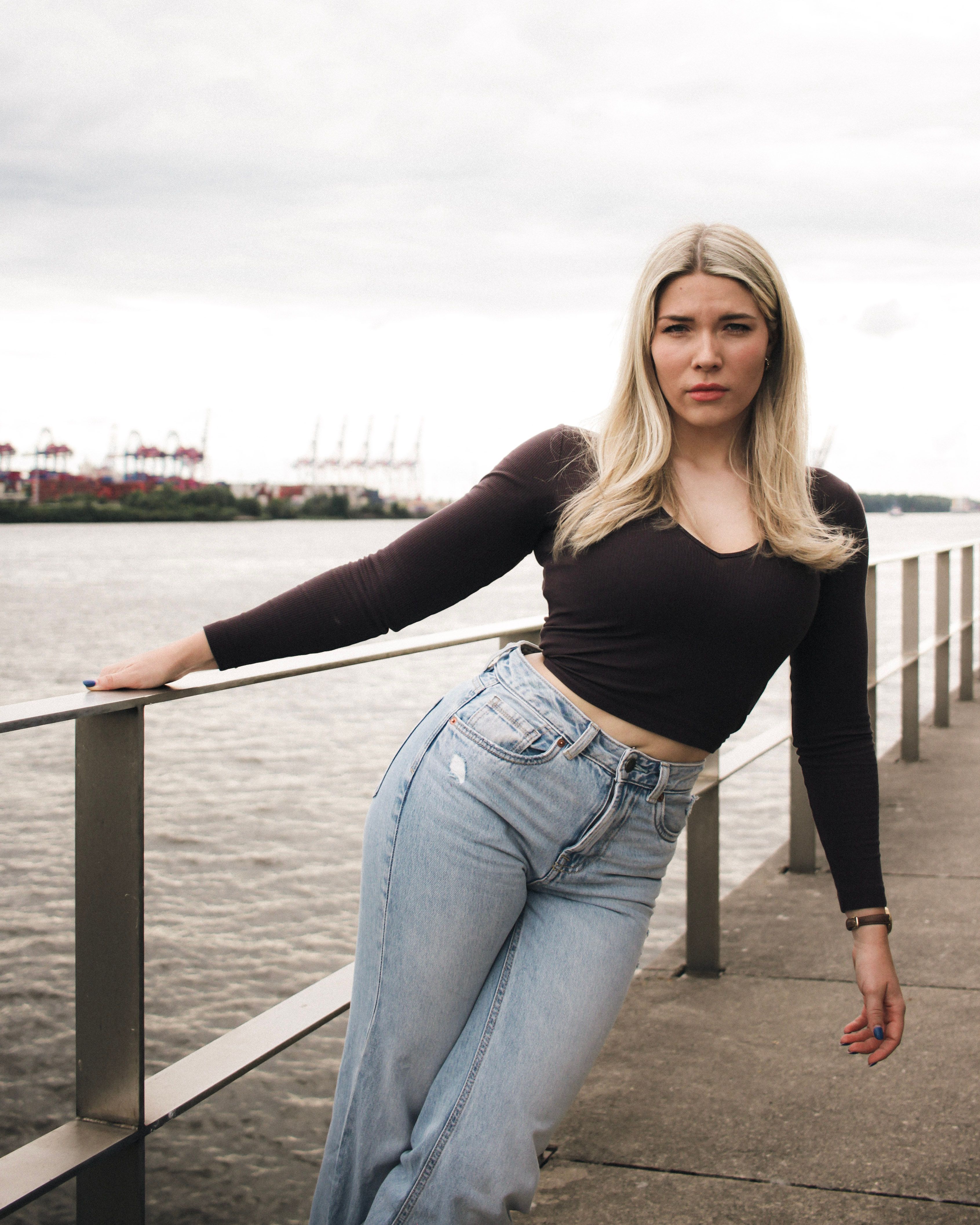 Young woman leans on railing by Elbe River, Hamburg, showing urban fashion and style.