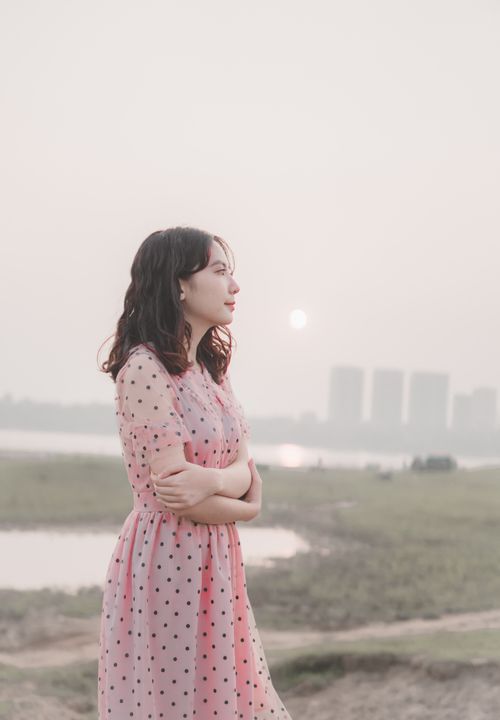 Free Side view of young contemplative ethnic female in dress standing with folded arms under sky with shiny sun and looking away Stock Photo