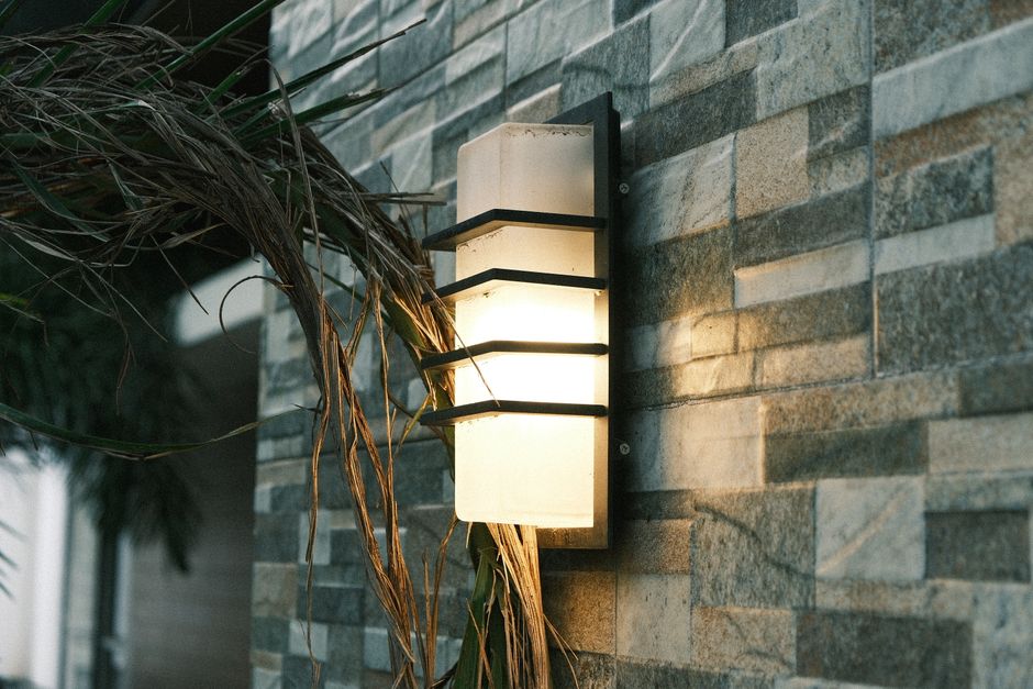 Illuminated outdoor wall lantern on a textured stone wall, surrounded by plants, setting a cozy evening ambiance.
