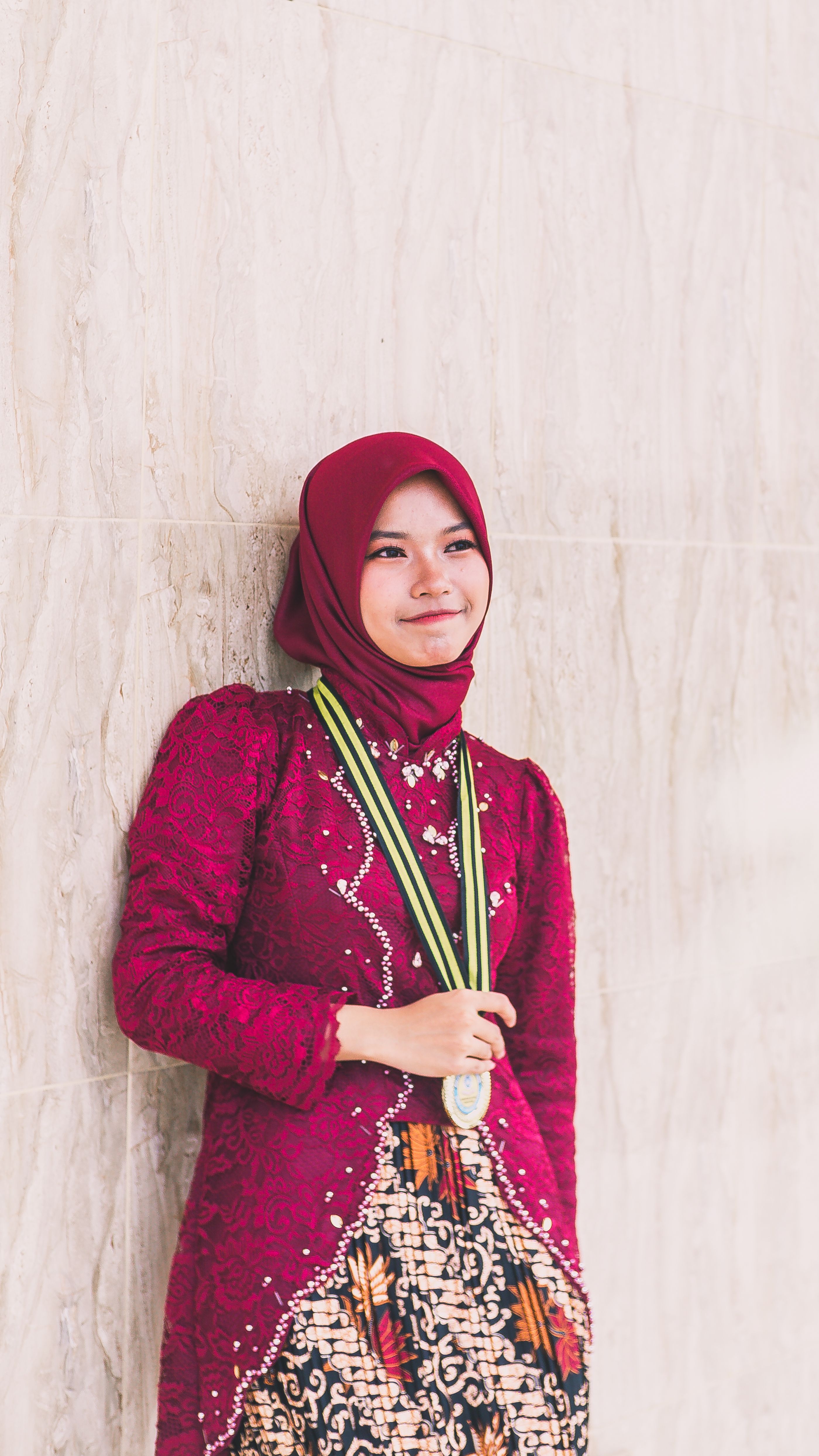 Smiling young woman in a red traditional dress and hijab, holding a medal, leaning against a marble wall.