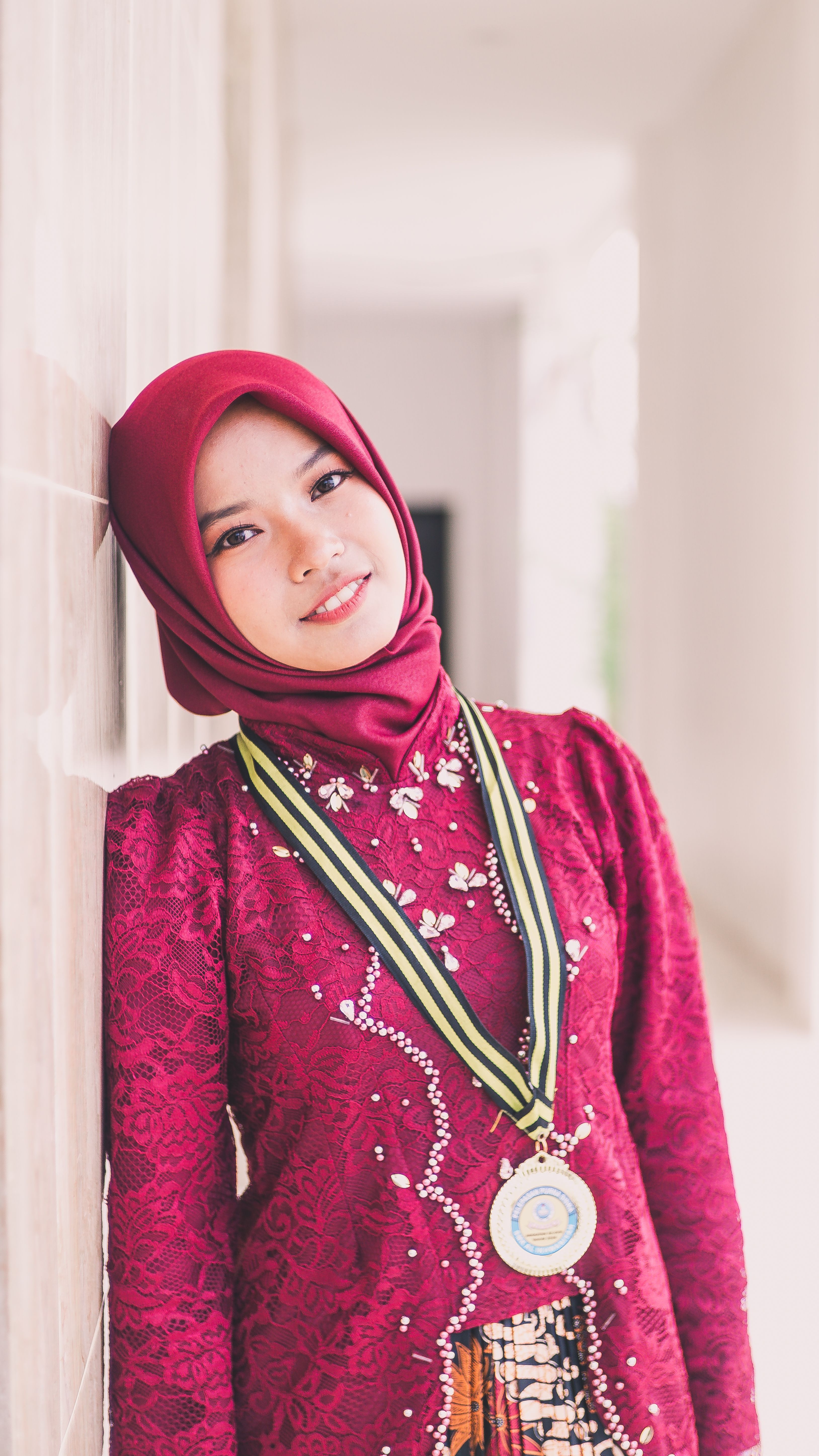 Smiling woman in red hijab and traditional dress with a medal, leaning against a wall.