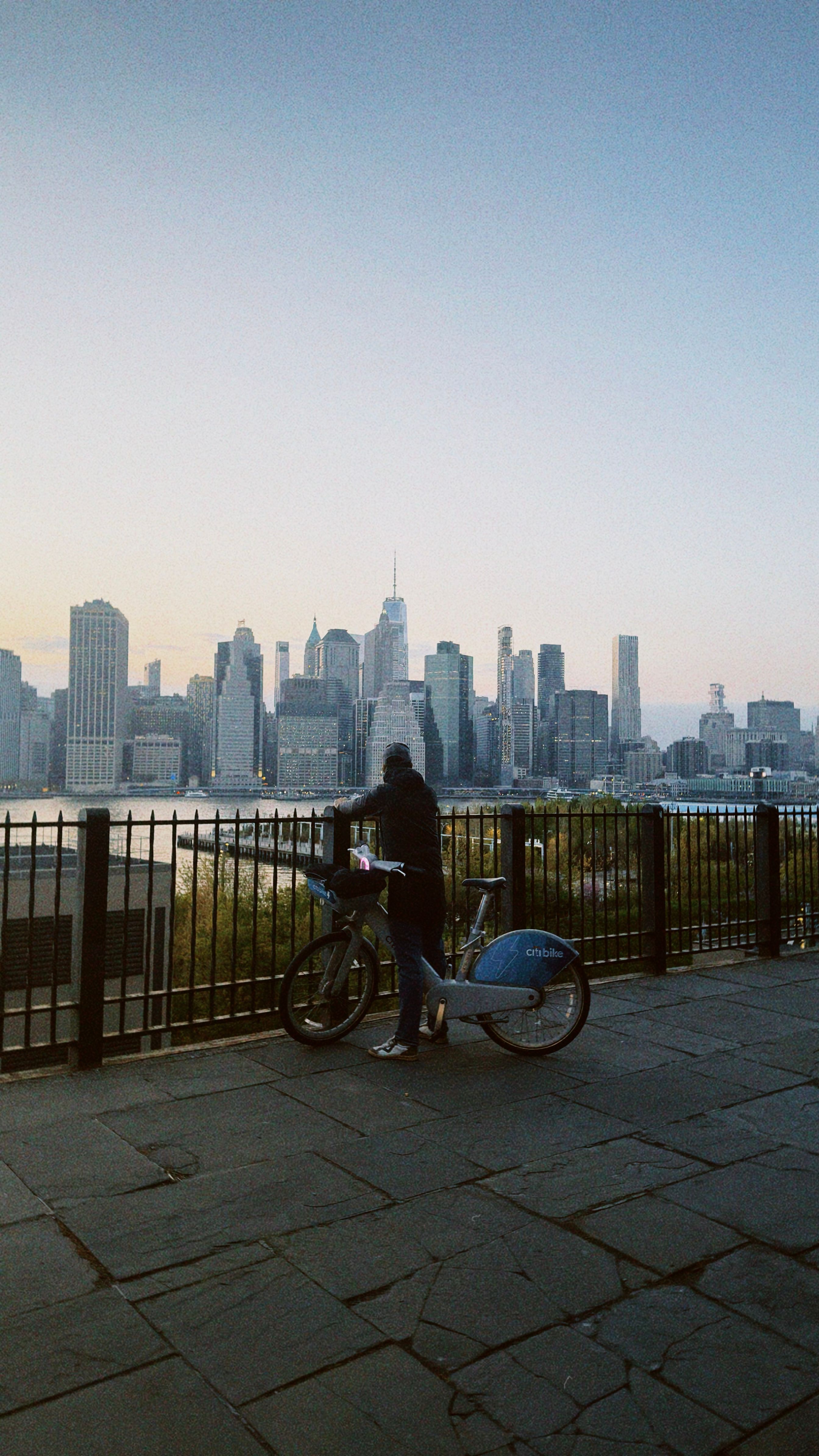 A person on a bike overlooks the New York City skyline from Brooklyn Heights during twilight.