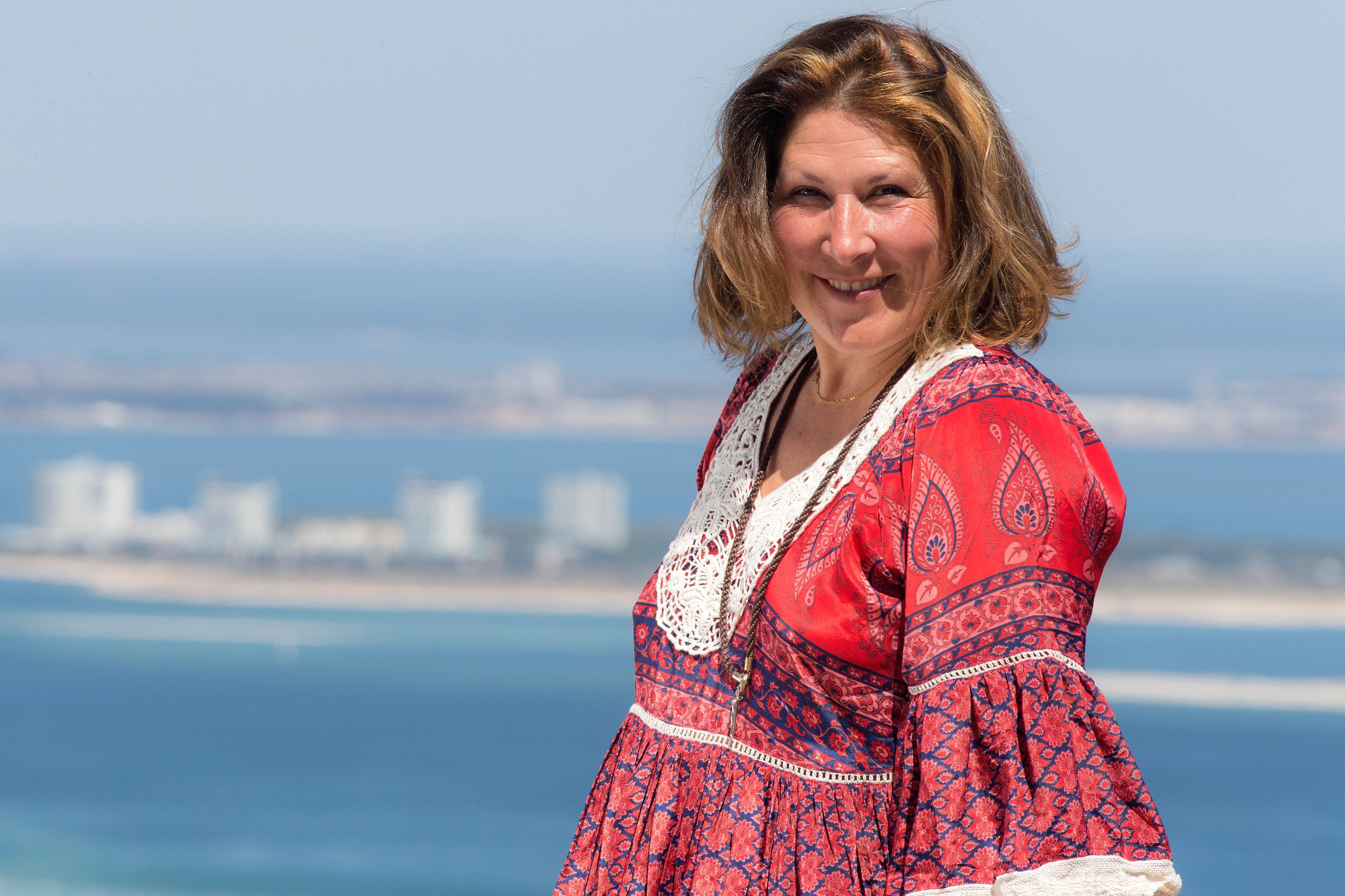 Smiling woman in a vibrant red dress enjoying a sunny day by the sea.