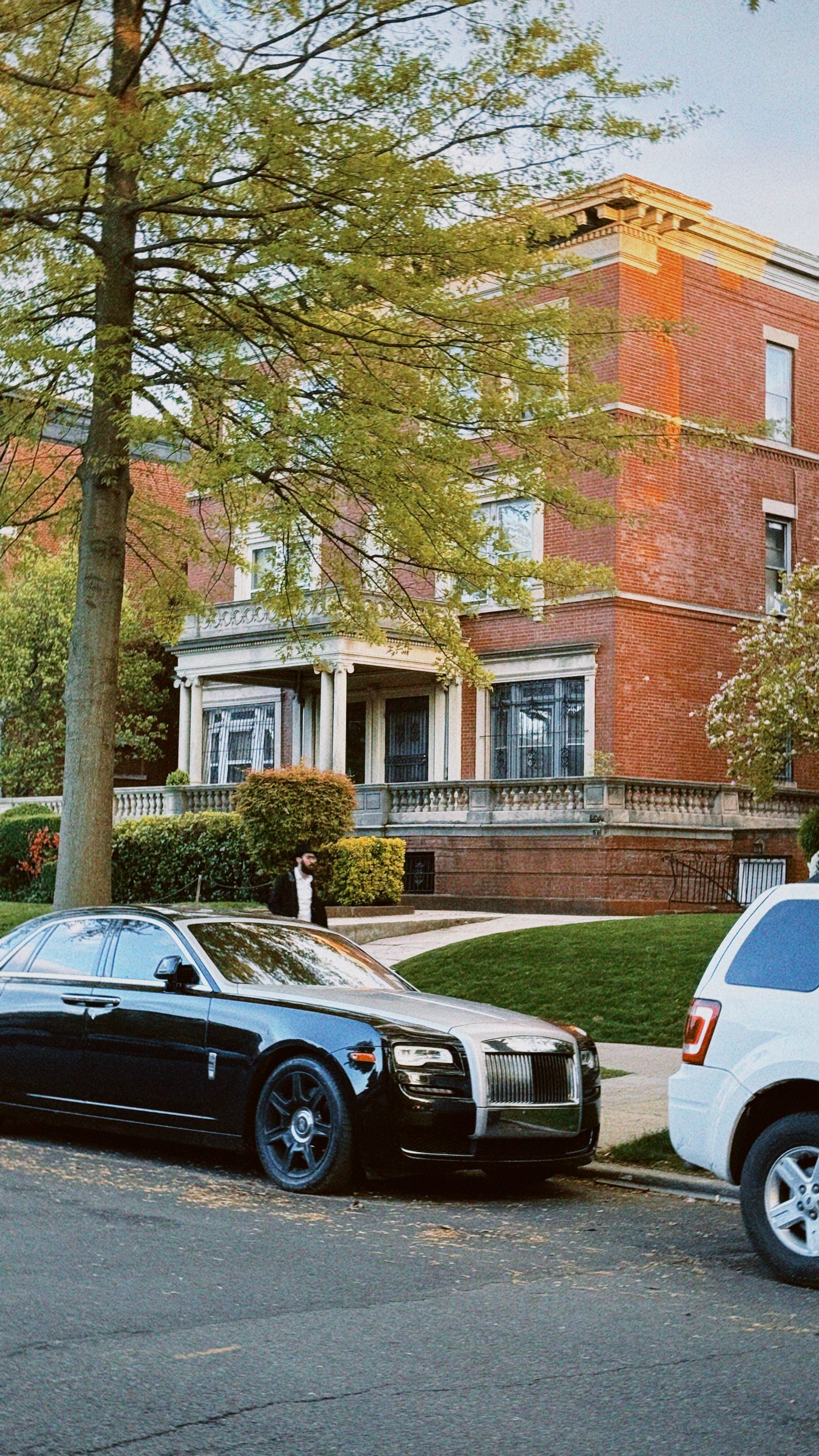 Elegant black luxury car parked on a street in front of a classic brick house surrounded by greenery.