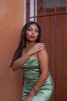 Elegant woman in a green satin dress poses by a rustic orange wall outdoors.