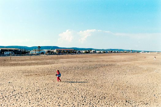 A solitary person walks on a vast, empty beach at Gruissan, Occitanie, France, under a clear blue sky.