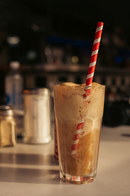 A tall glass mug of root beer float with a scoop of vanilla ice cream floating in fizzy root beer, topped with whipped cream and a cherry, with a straw and long spoon resting in the glass.