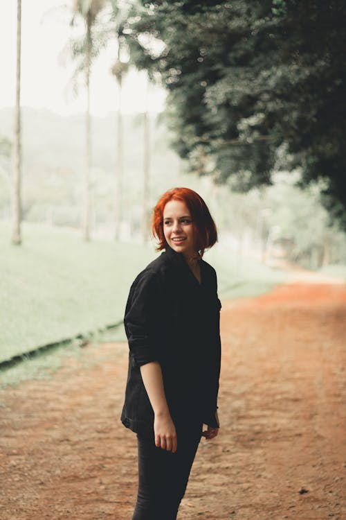 Side view of smiling redhead female in black hoodie standing on pathway in park looking away