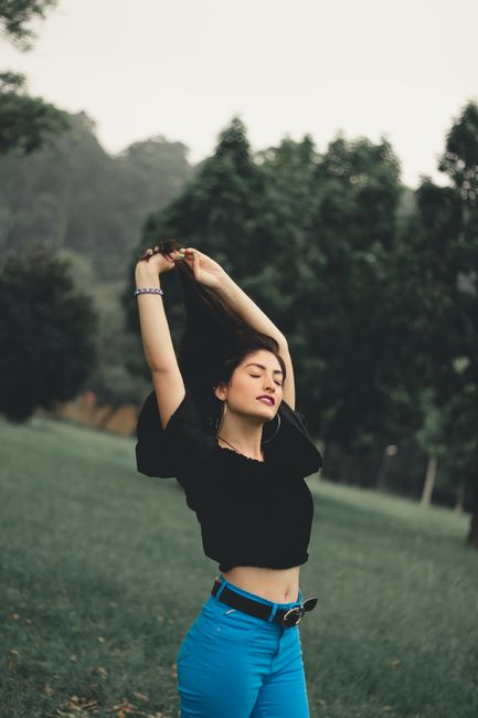 Calm slender ethnic woman in denim and black top stretching arms up closing eyes in leisure standing in countryside