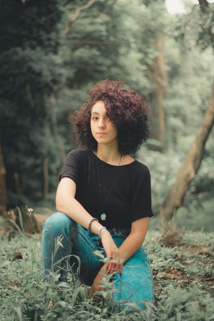 Young ethnic female with curls looking at camera while sitting on green lawn in peaceful nature