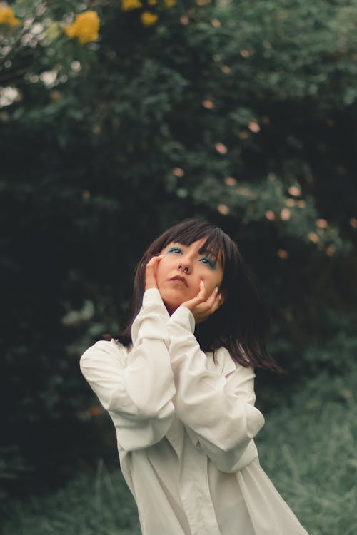 Young dreamy Asian woman in white shirt touching cheeks while looking up against green trees