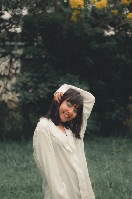 Happy young Asian female in white shirt touching hair with eyes closed laughing against green trees