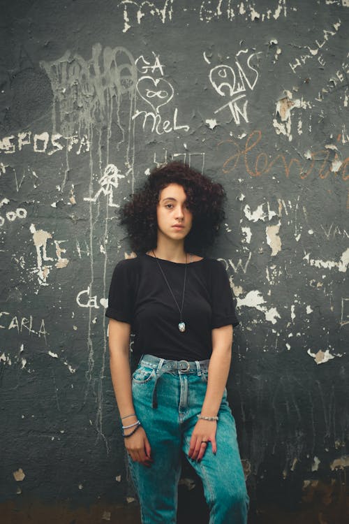 Woman in Black Crew Neck T-shirt and Blue Denim Pants Standing Beside Vandalized Wall