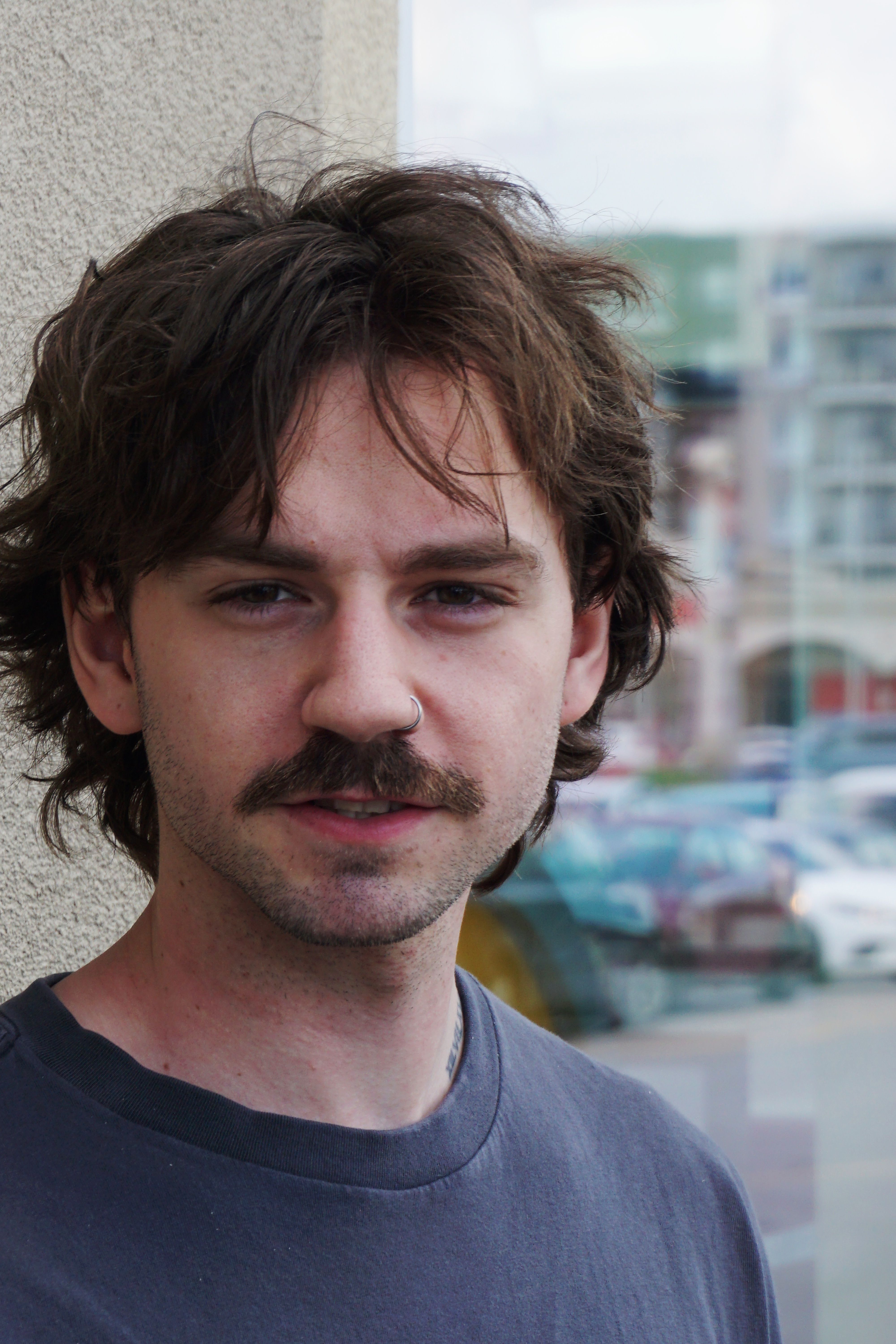 Close-up portrait of a young man with a mustache standing outdoors against a city backdrop.