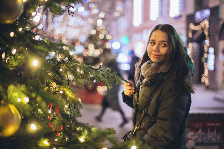 Woman In Bubble Jacket Standing Near Christmas Tree With String Lights