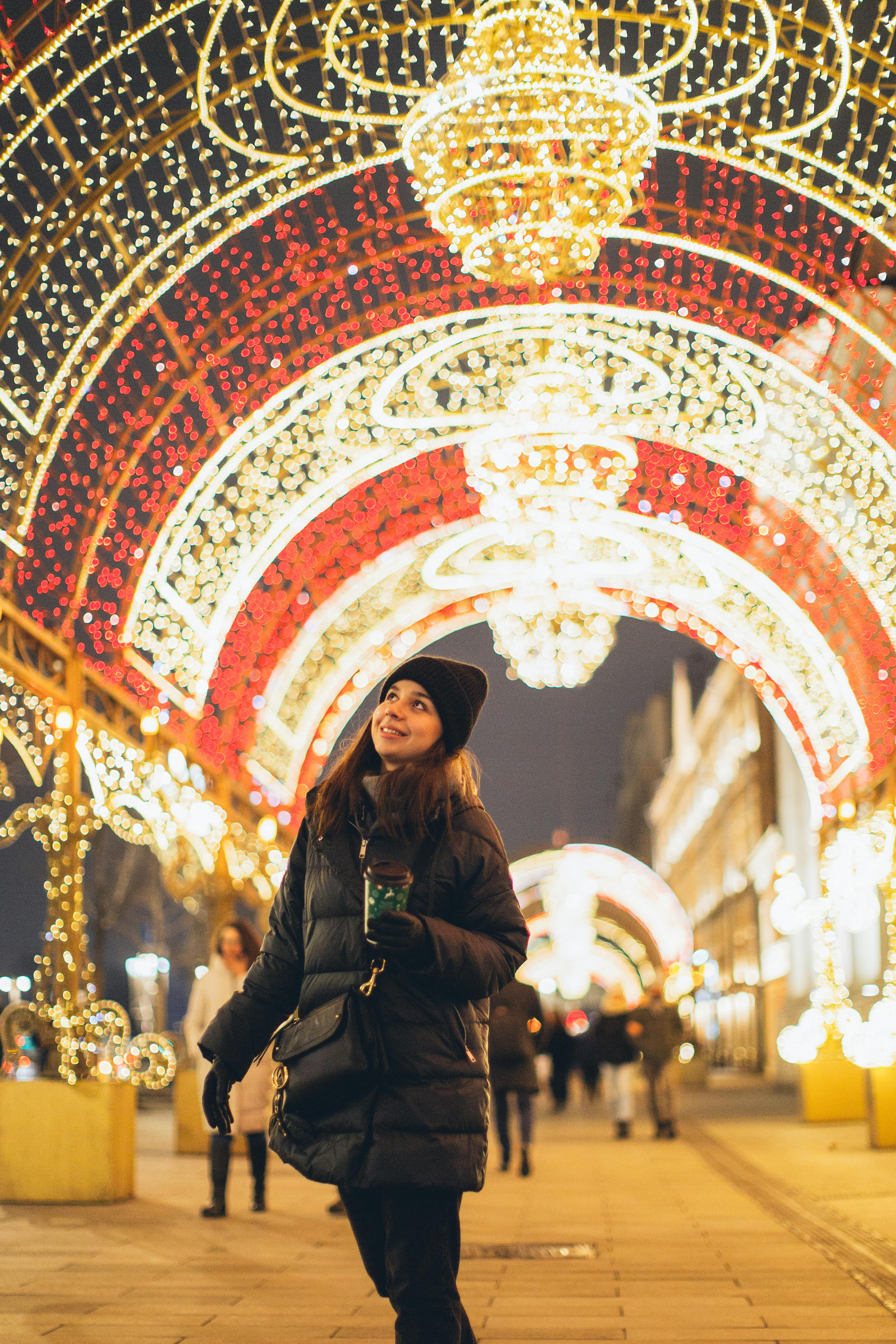 woman in black jacket standing against lighted christmas lights arch