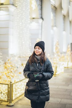 一个戴着无檐帽、穿着冬季外套的女人，面带微笑，手拿咖啡，走在节日灯光装饰的街道上