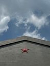 A modern concrete facade with a prominent red star, set against a vibrant blue sky with clouds.