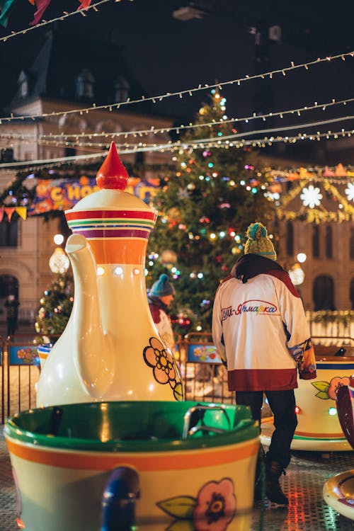 Employee servicing carousel walking among giant teapot and cups in evening park decorated in Christmas style
