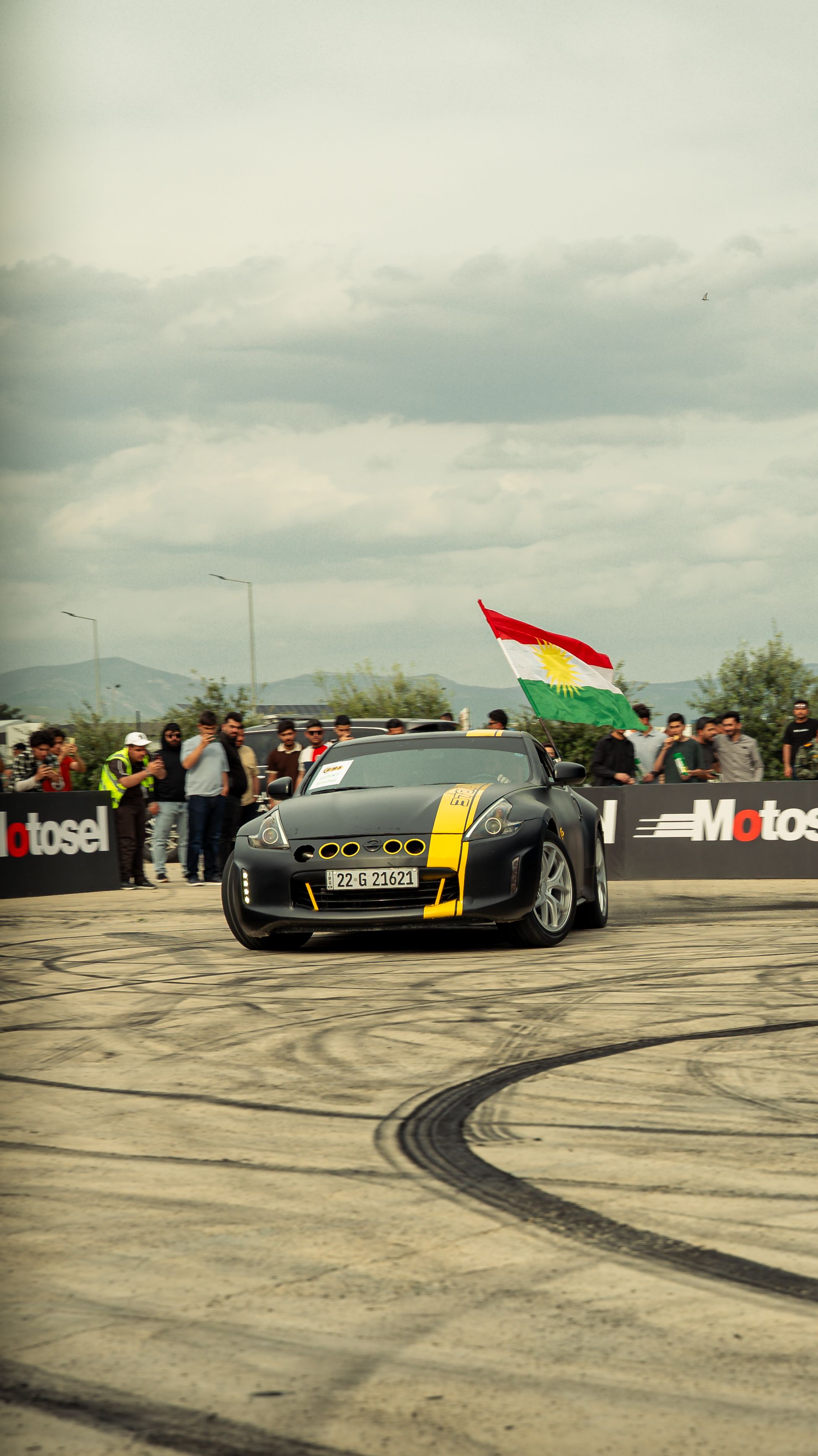 Dynamic car drifting with Kurdistan flag in Sulaymaniyah. Enthusiastic crowd and cloudy skies.