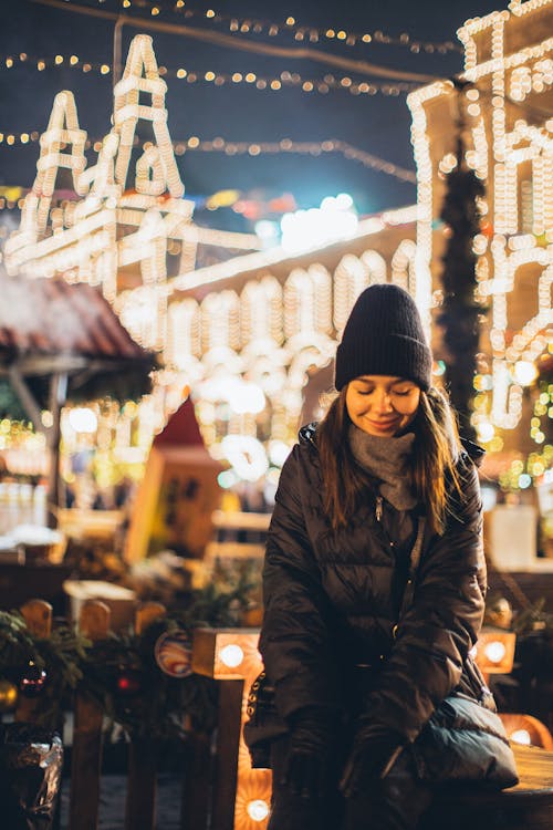 Happy young female in black warm jacket and hat looking down and smiling while sitting on wooden bench among Christmas decorations in night city