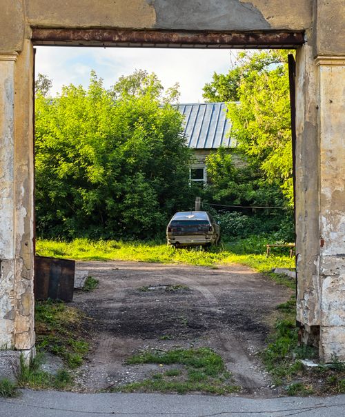Free A rustic gateway frames an abandoned car surrounded by lush greenery, highlighting nature's reclamation of urban spaces. Stock Photo