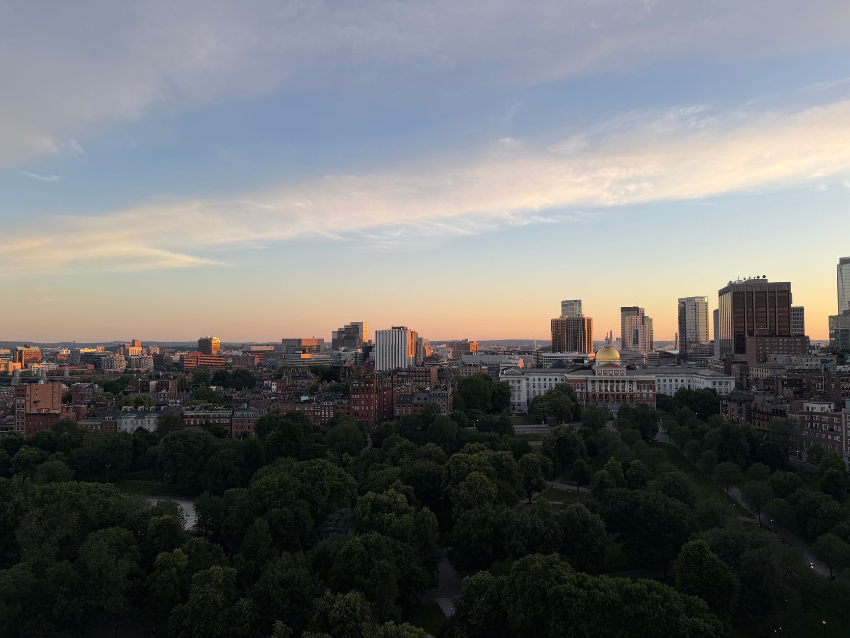 Aerial view of Boston skyline at sunset, featuring lush greenery and city buildings.