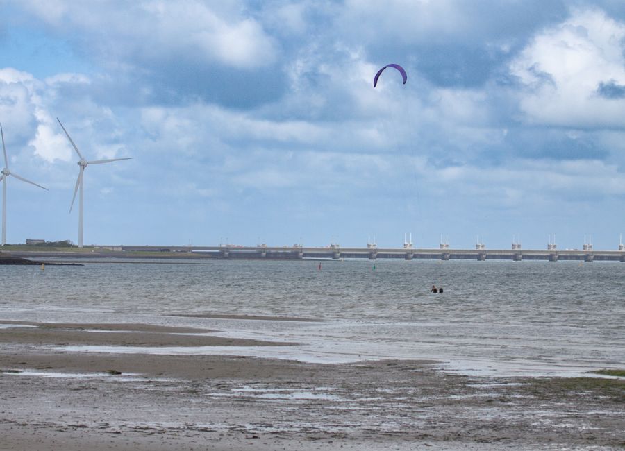 A scenic beach view at Kamperland with wind turbines and a kite surfer under a cloudy sky.
