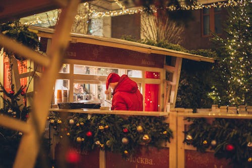 Back view of male wearing warm outerwear standing near counter at Christmas fair while enjoying holidays and purchasing takeaway beverage in illuminated city
