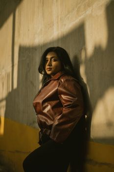 A woman leans against a sunlit wall, wearing a brown leather jacket.