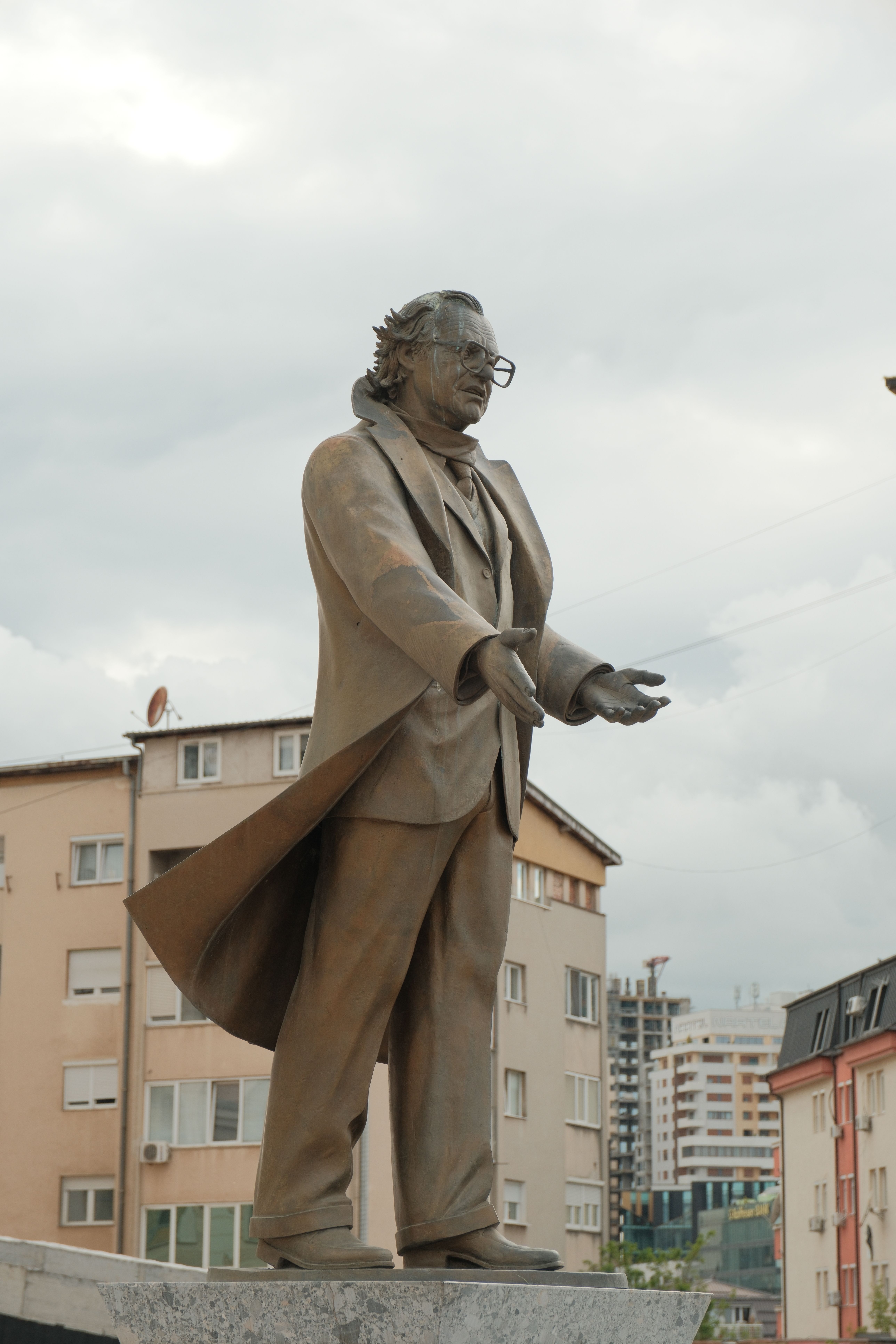Bronze statue of a man in a suit, set against city buildings in Priştina.