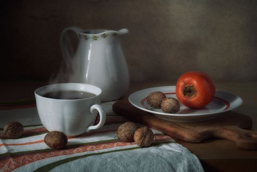 Warm still life scene featuring a cup of tea, a jug, nuts, and persimmon on a wooden board.