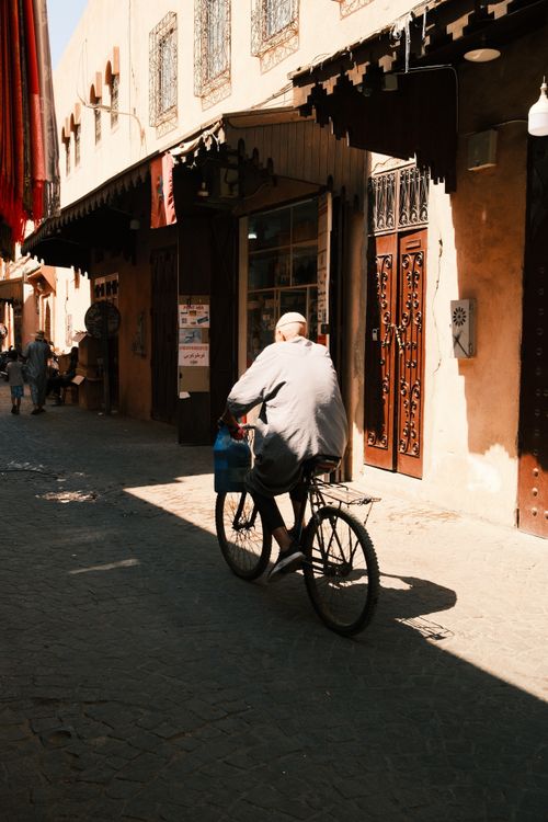 Free A man on a bicycle rides through a sunlit alley in a historic city, showcasing traditional architecture. Stock Photo