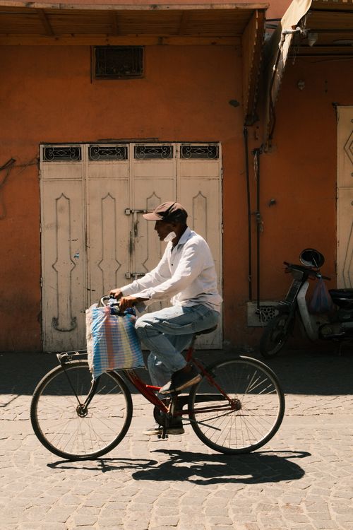 Free A man rides a bicycle in a sunlit urban alley, casting shadows on the cobblestone street. Stock Photo