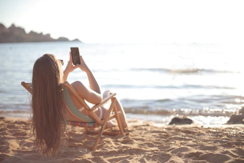 免費 白色背心的女人坐在海灘上的白色木椅上 圖庫相片