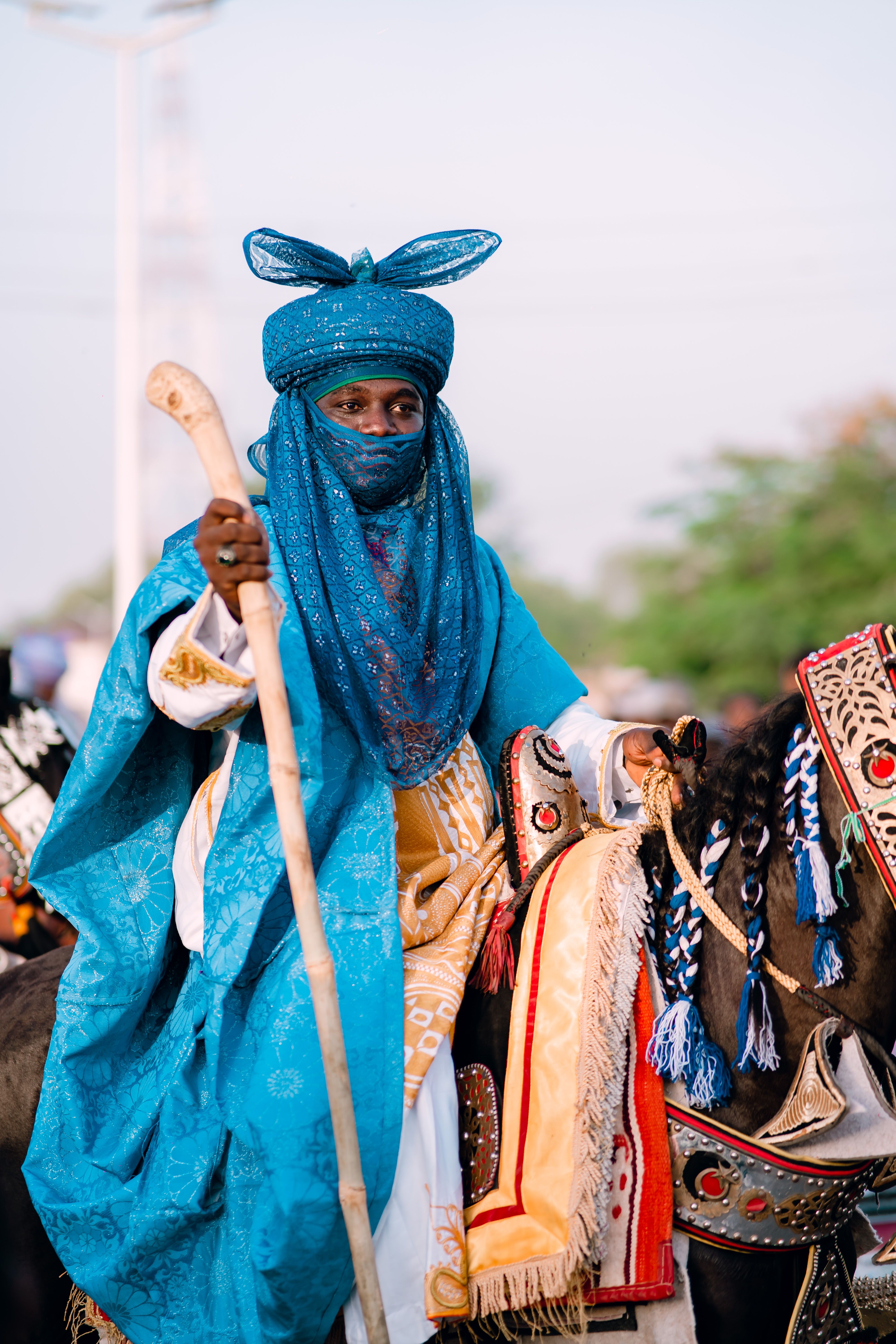 A Tuareg man in vibrant blue attire rides a decorated horse at a cultural event.