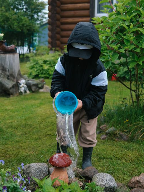 Free A child wearing a hood pours water from a blue bucket in a garden with greenery. Stock Photo
