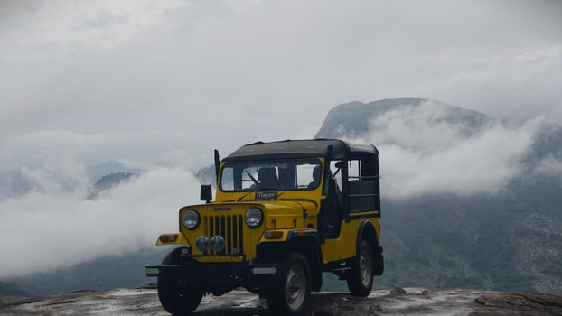 A yellow jeep perched on a mountain cliff with a misty, cloud-covered background.