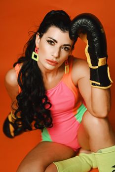 Energetic woman in vibrant fitness attire with boxing gloves on vibrant orange background.