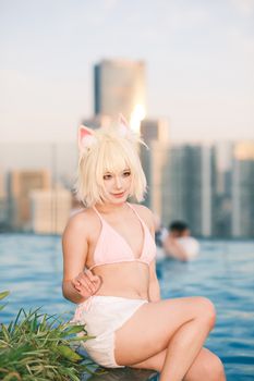 Blond woman in anime cosplay costume relaxing by rooftop pool with city skyline backdrop.