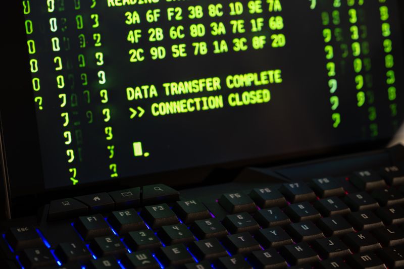 A laptop terminal screen showing hexadecimal data and a "DATA TRANSFER COMPLETE" message, representing the cyber operation infrastructure